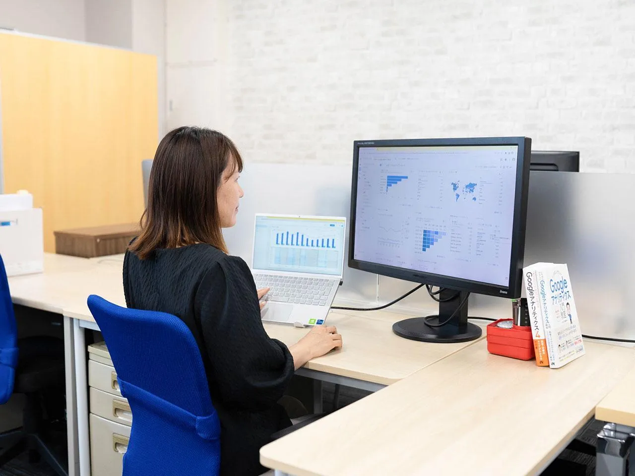 A businesswoman sitting at her office desk, analyzing web data on a large monitor alongside her laptop, with a Google Analytics book nearby.
