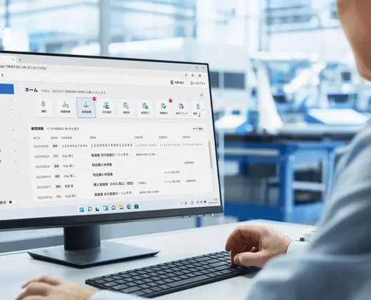 Over-the-shoulder view of a person using a desktop computer to navigate a Japanese business management software dashboard, set in a bright manufacturing or industrial environment.
