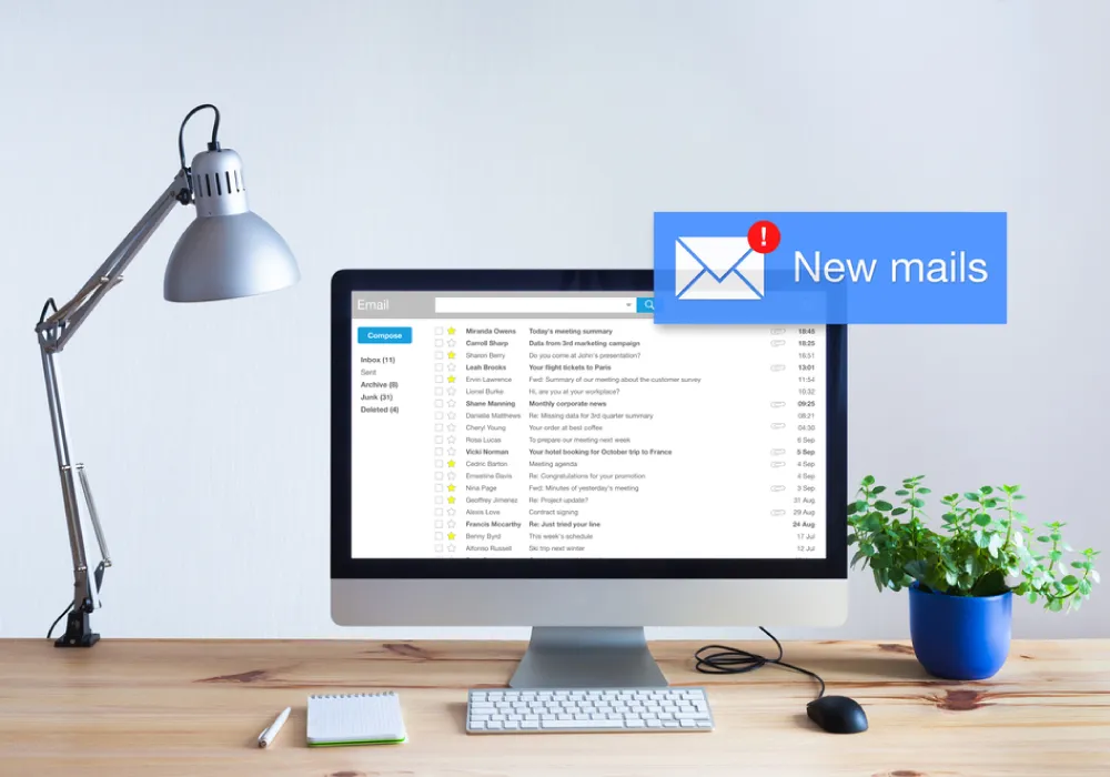 A clean wooden desk setup featuring a desktop computer displaying an email inbox with a prominent 'New mails' notification popup, flanked by a desk lamp and a potted plant.