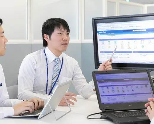 Three businessmen in an office collaborating over a software application, with one man pointing a pen at a desktop monitor displaying a dashboard interface while another looks at a laptop.