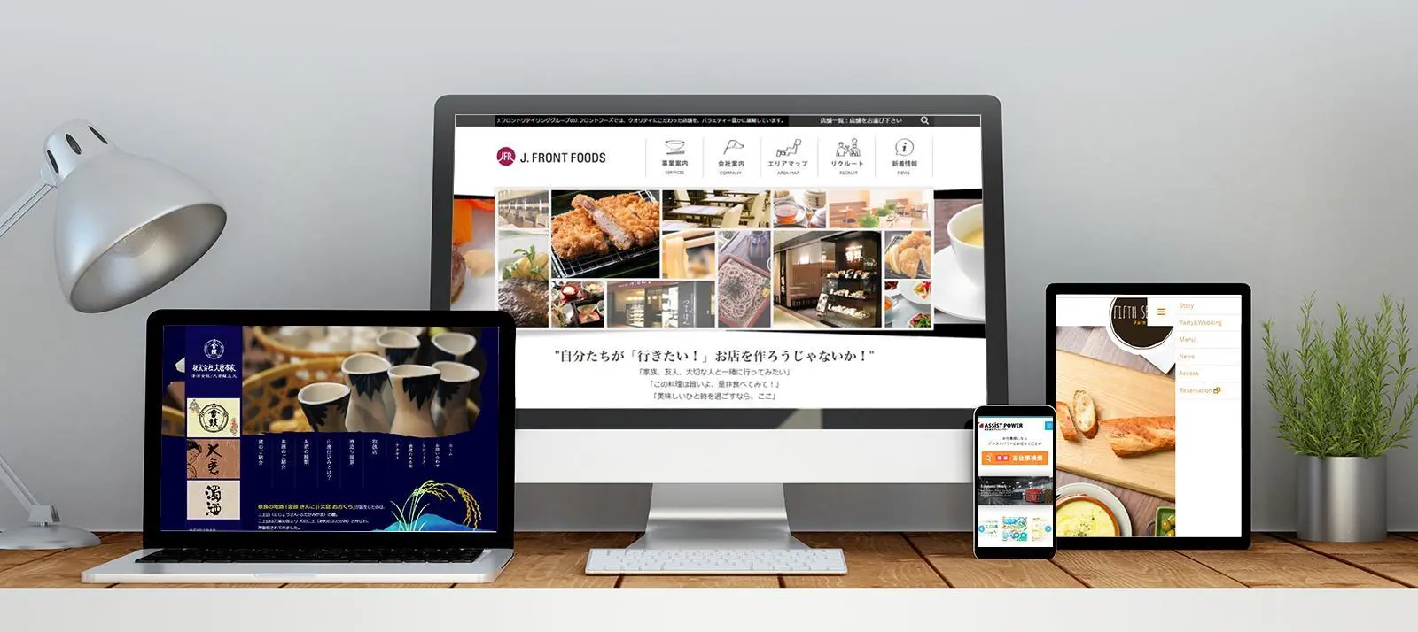 A desktop monitor, laptop, tablet, and smartphone displaying different responsive layouts of restaurant websites, arranged on a wooden desk next to a lamp and a potted plant.