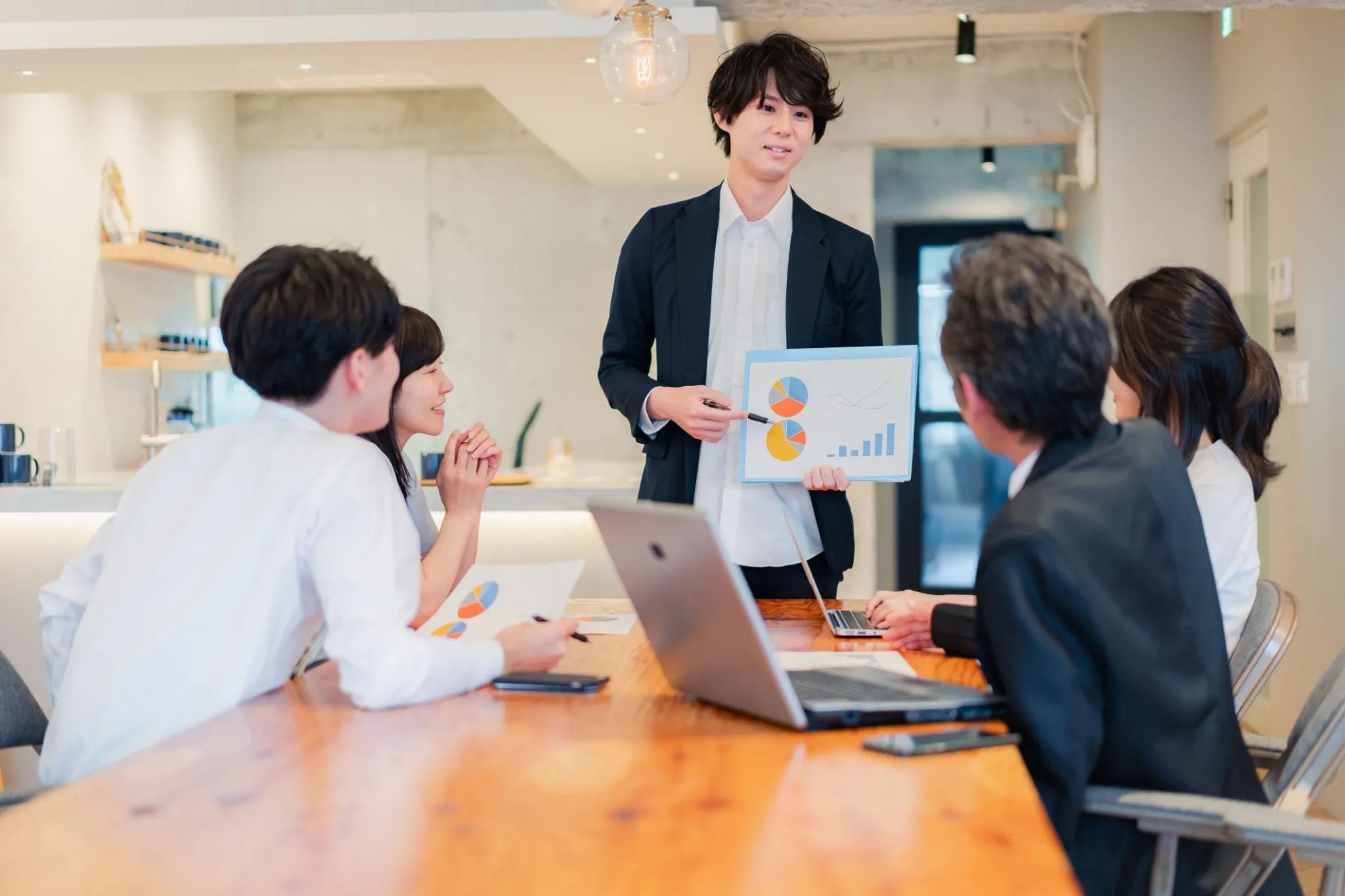 A young businessman presenting a document with data charts to his seated team members during a meeting around a wooden conference table.