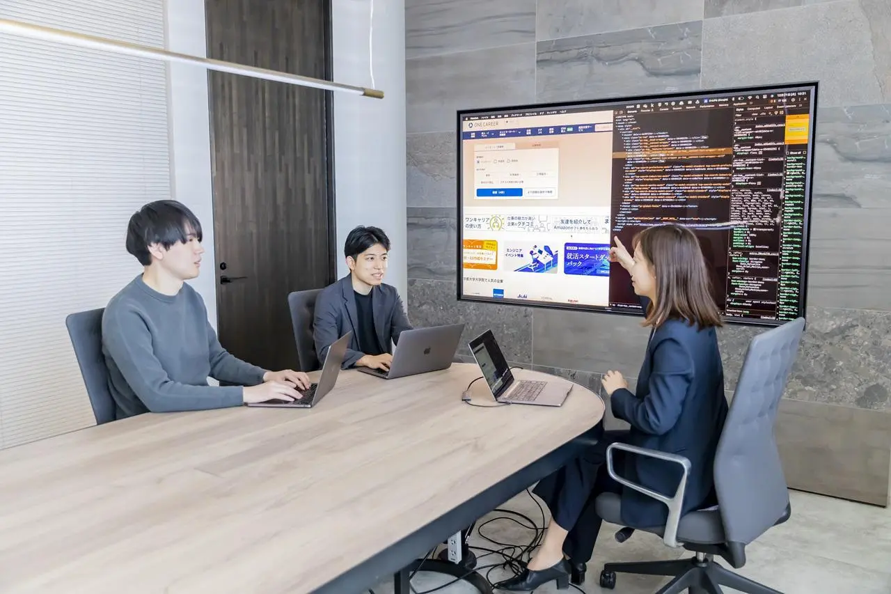 Three colleagues in a modern conference room discussing a project, with a woman pointing at a large wall monitor that displays a website layout alongside its HTML/CSS code.